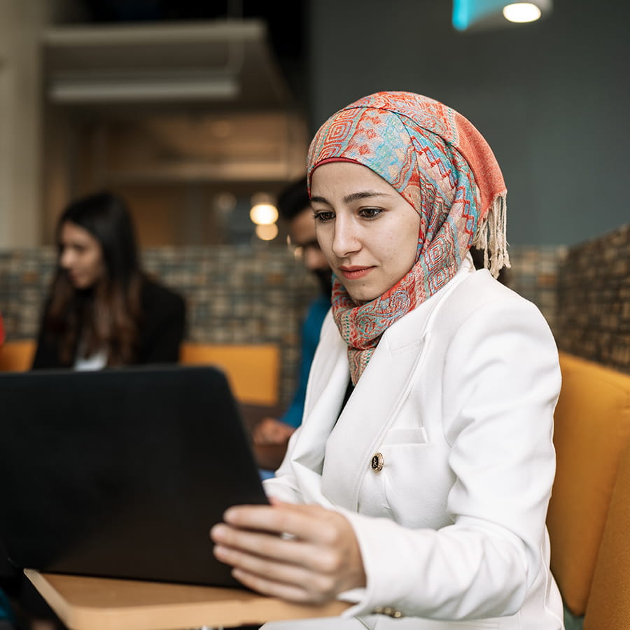Student, Abrar Ahmed, in white blazer and patterned headscarf looking at laptop.