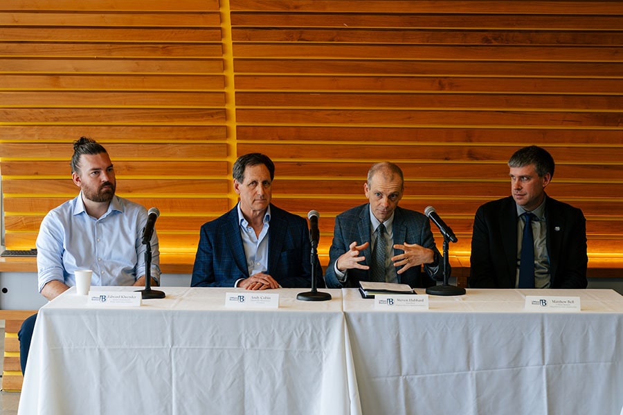The four panelists of the quantum roundtable discussion sit at a table covered in a white tablecloth in front of a wooden wall.